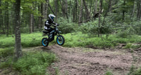 Rider jumping a blue Buzymoto electric dirt bike on a forest trail, featured by Dirt Bike Mall