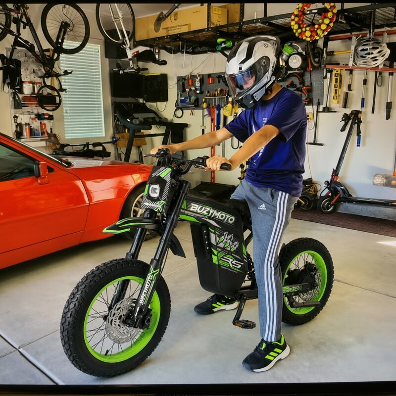 Young rider in helmet sitting on Buzymoto DAT70 green electric dirt bike inside a home garage