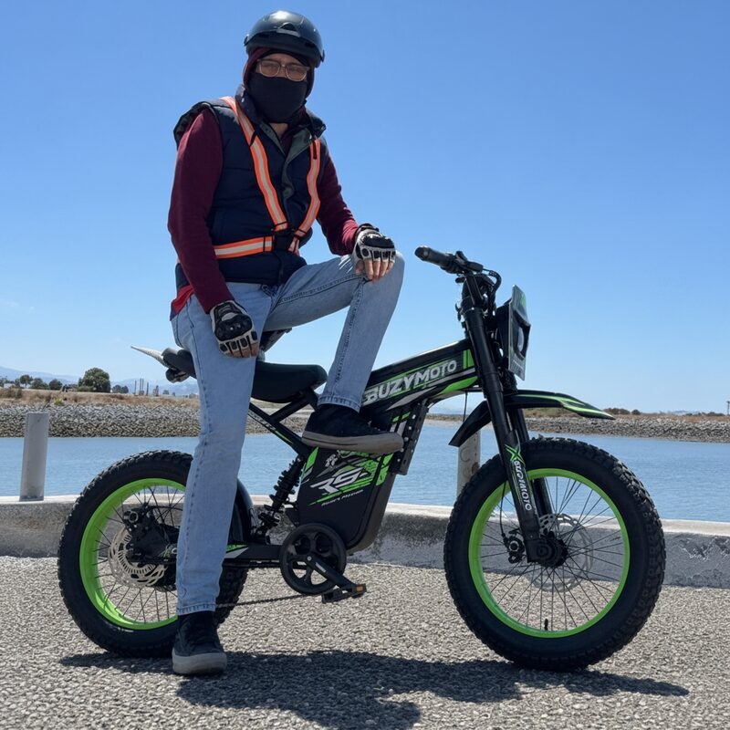Rider posing on Buzymoto DAT70 green electric dirt bike at a sunny waterfront location