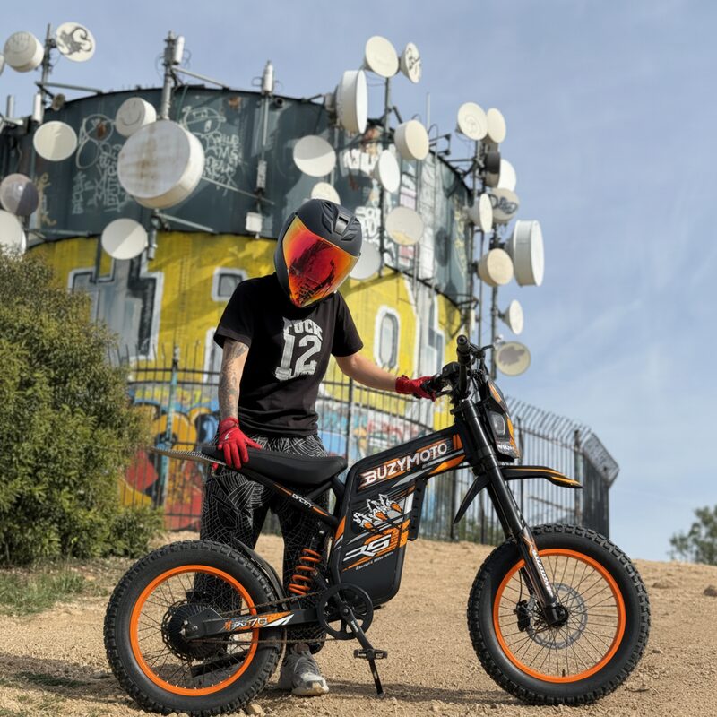 Rider in helmet posing with Buzymoto DAT70 orange electric dirt bike in front of urban graffiti wall