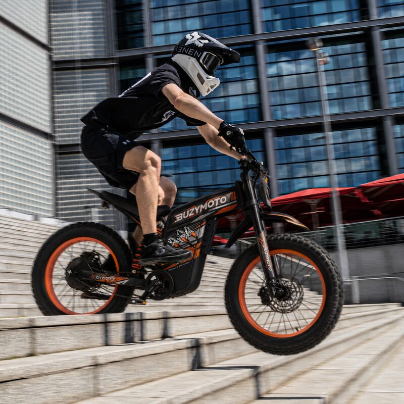 Rider performing stunt on Buzymoto DAT70 orange electric dirt bike jumping down urban stairs