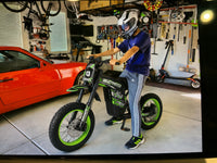 Rider sitting on a green Buzymoto electric dirt bike inside a garage, featured by Dirt Bike Mall