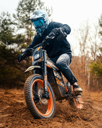 Rider taking an orange Buzymoto electric dirt bike along a rugged trail from Dirt Bike Mall