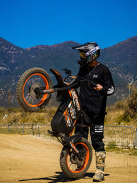 Rider performing a mountain wheelie on an orange Buzymoto electric dirt bike from Dirt Bike Mall