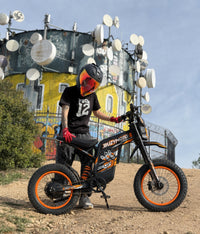 Orange Buzymoto electric dirt bike on an outdoor trail near a tower