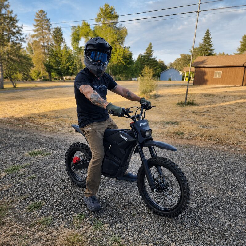 Adult rider in helmet straddling Terrawulf M7 black electric dirt bike on gravel road at sunset